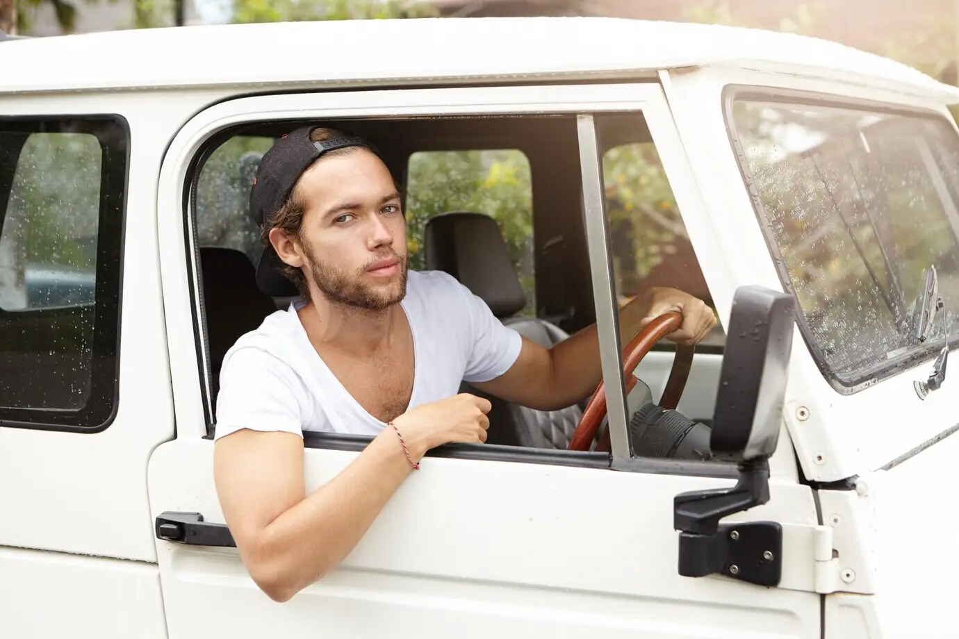 Gros plan d’un jeune mannequin barbu, beau et stylé, posant à l’intérieur d’une jeep blanche, assis au siège conducteur, une main posée sur le volant et affichant une expression confiante sur le visage.